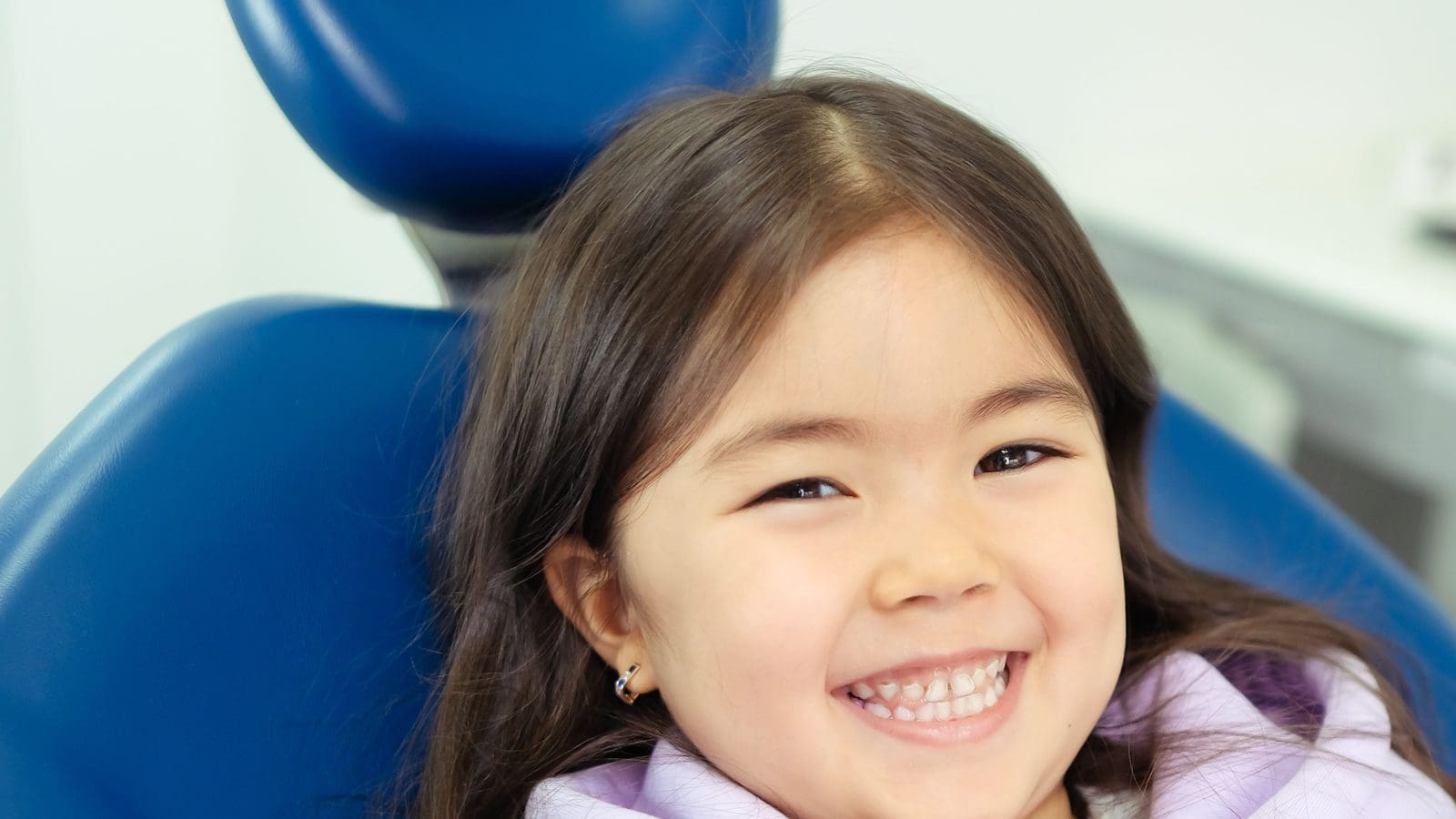 Young girl grinning in a blue dental chair at a check on the best age for braces