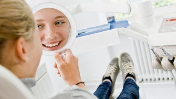 Teen smiling at her reflection in a hand mirror in the orthodontist's chair, the moment braces come off