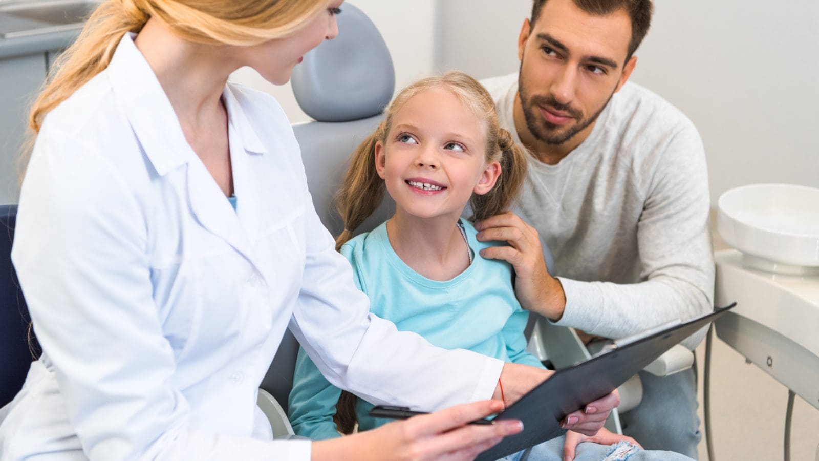 Girl in the dental chair with her dad as the orthodontist reviews her chart, choosing the best orthodontist