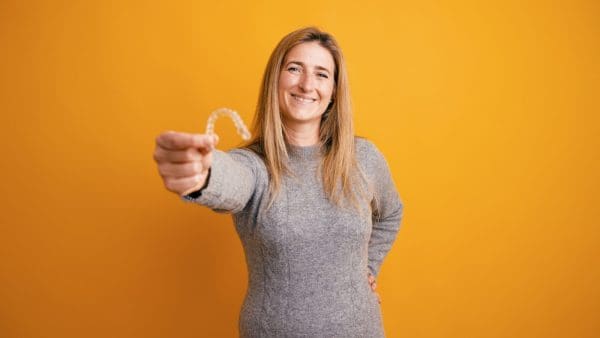 Smiling woman holding up a clear aligner against a bright yellow wall, the tray behind clear aligner cost