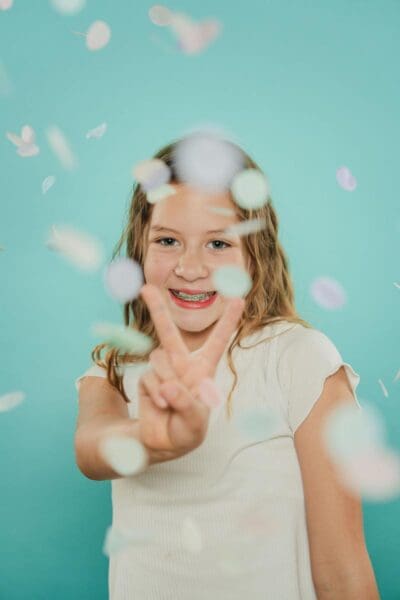 tooth-by-tooth-orthodontics-tooth-by-tooth-dr-patel-home-slider-girl-patient-braces-peace-sign-confetti