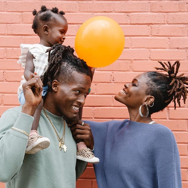 Family enjoying moment together against coral wall, child on parent's shoulders with orange balloon at Tooth by Tooth